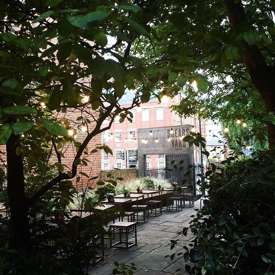 Outdoor seating area at Shears Yard with wooden tables and chairs, surrounded by lush green foliage, string lights, and a view of the red-brick building facade with the Shears Yard sign.