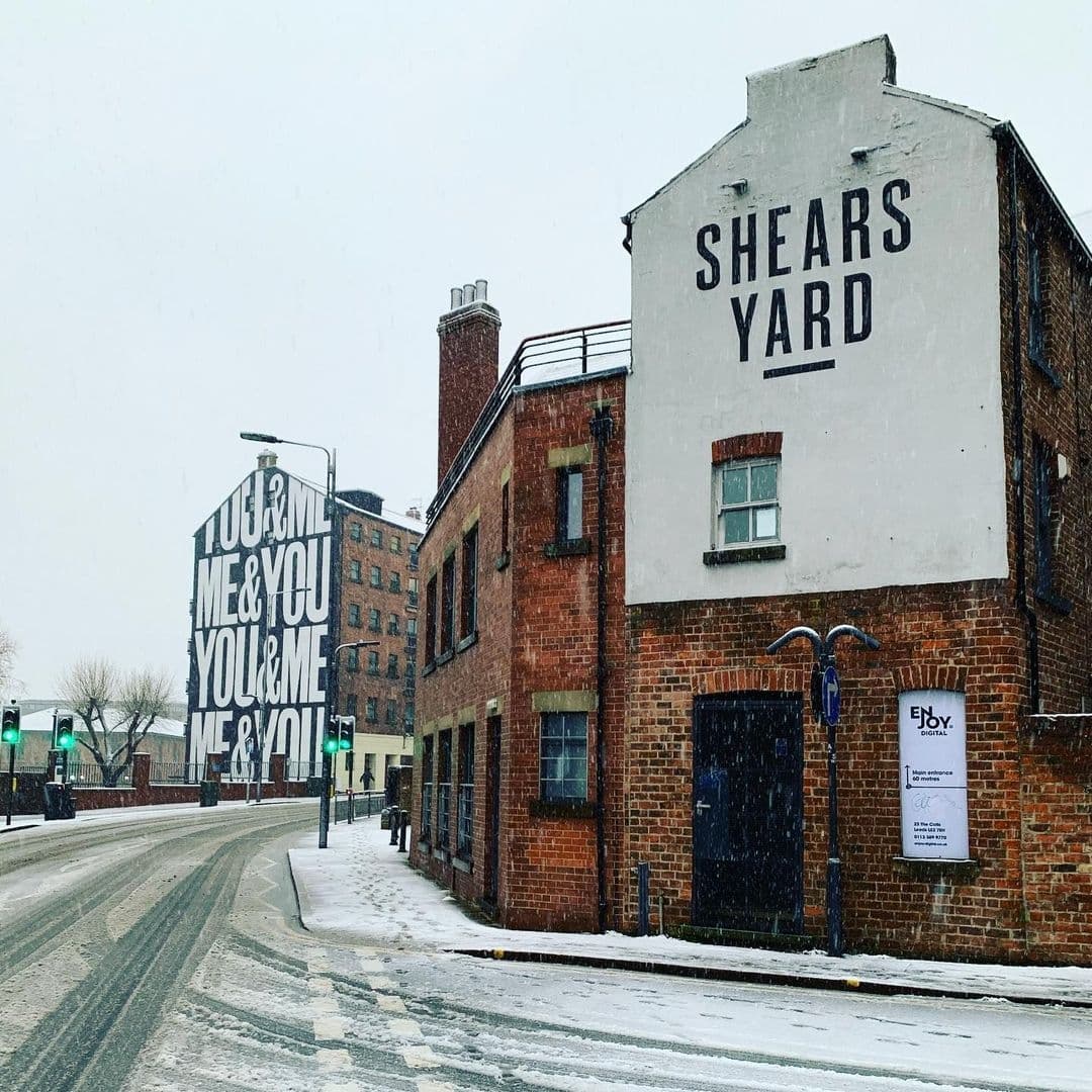 "Winter scene at Shears Yard with snow-covered streets, a brick building prominently displaying the 'Shears Yard' sign, and another building in the background with a large 'You & Me' mural."