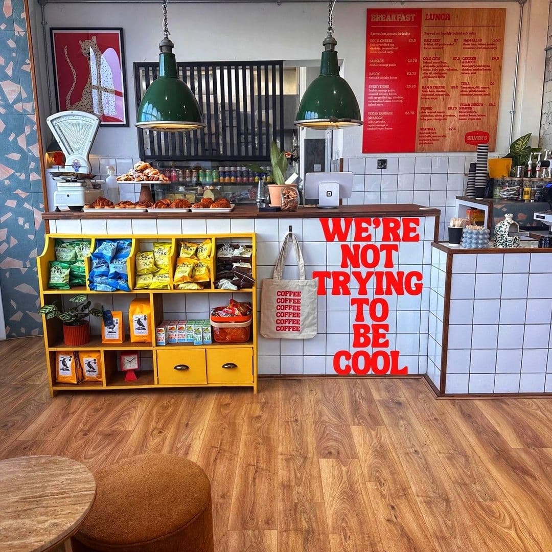 Interior of Silver’s Deli featuring a vibrant yellow shelf with snacks, a counter with pastries, green pendant lights, and a wall with the text "We're not trying to be cool."