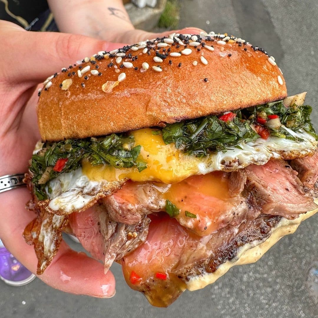 Close-up of a gourmet sandwich from Silver’s Deli featuring a sesame seed bun, juicy steak, melted cheese, greens, and sauce, held in a person's hand.
