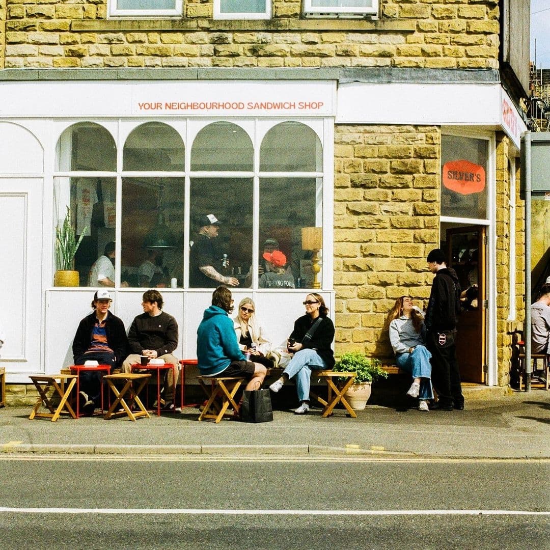 People enjoying food and drinks outside Silver's Deli, a neighborhood sandwich shop with a stone facade and large windows.
