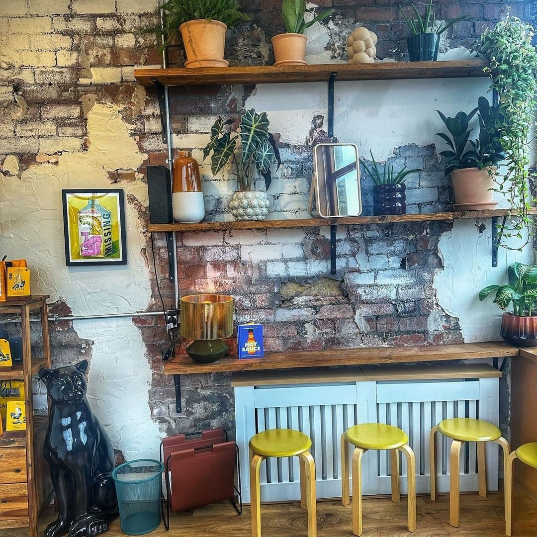 Interior corner of Silver's Deli featuring rustic exposed brick walls with white paint, wooden shelves with plants and decor, a black cat figurine, and yellow stools under a wooden counter.