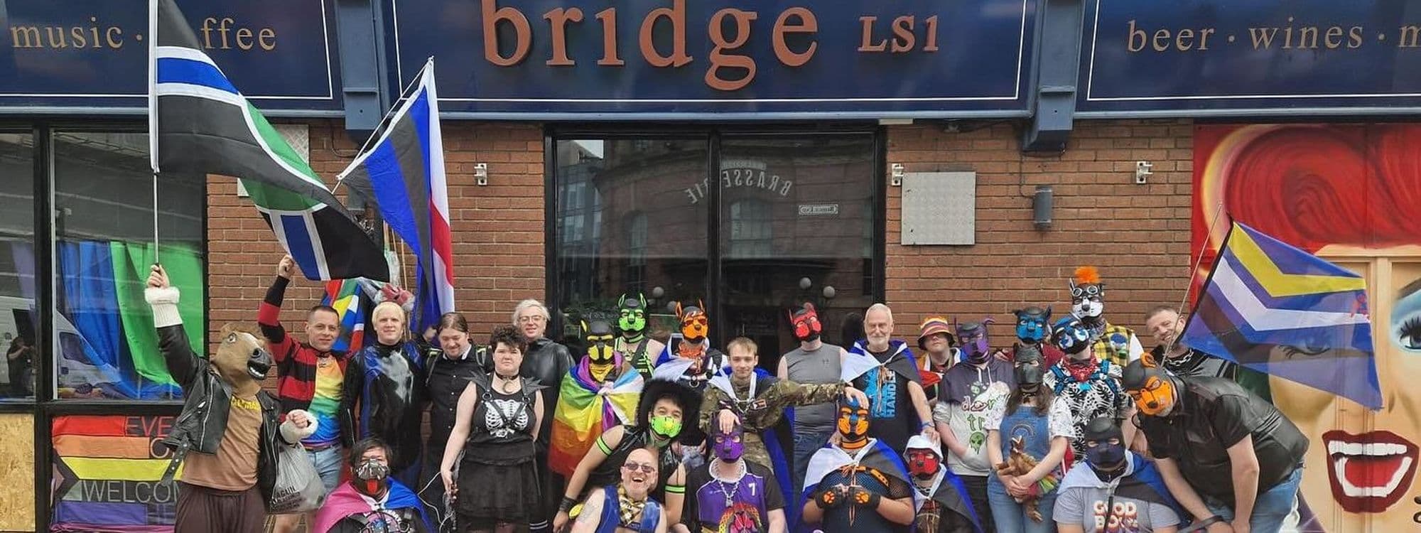 Group of people in colorful costumes and masks standing in front of The Bridge Bar LS1, with pride flags and a vibrant atmosphere.