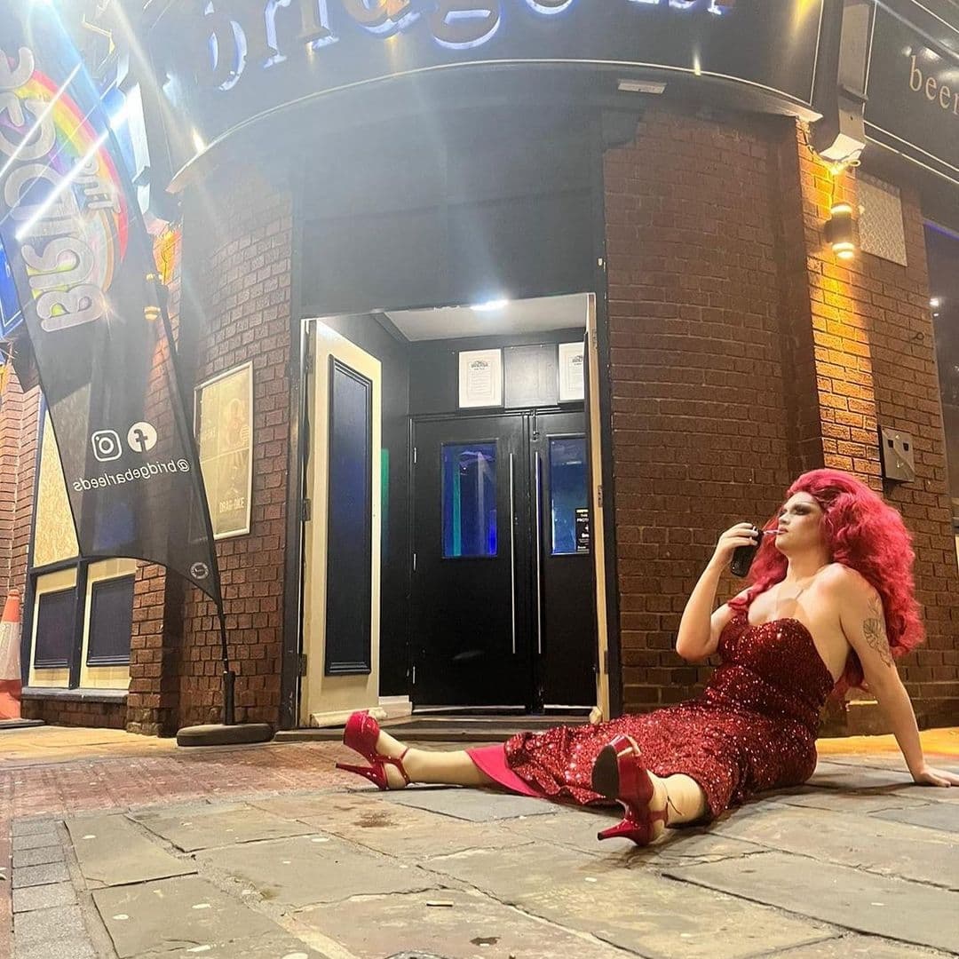 Person in a red dress and red shoes with red hair sitting on the pavement outside The Bridge Bar at night, looking thoughtful.
