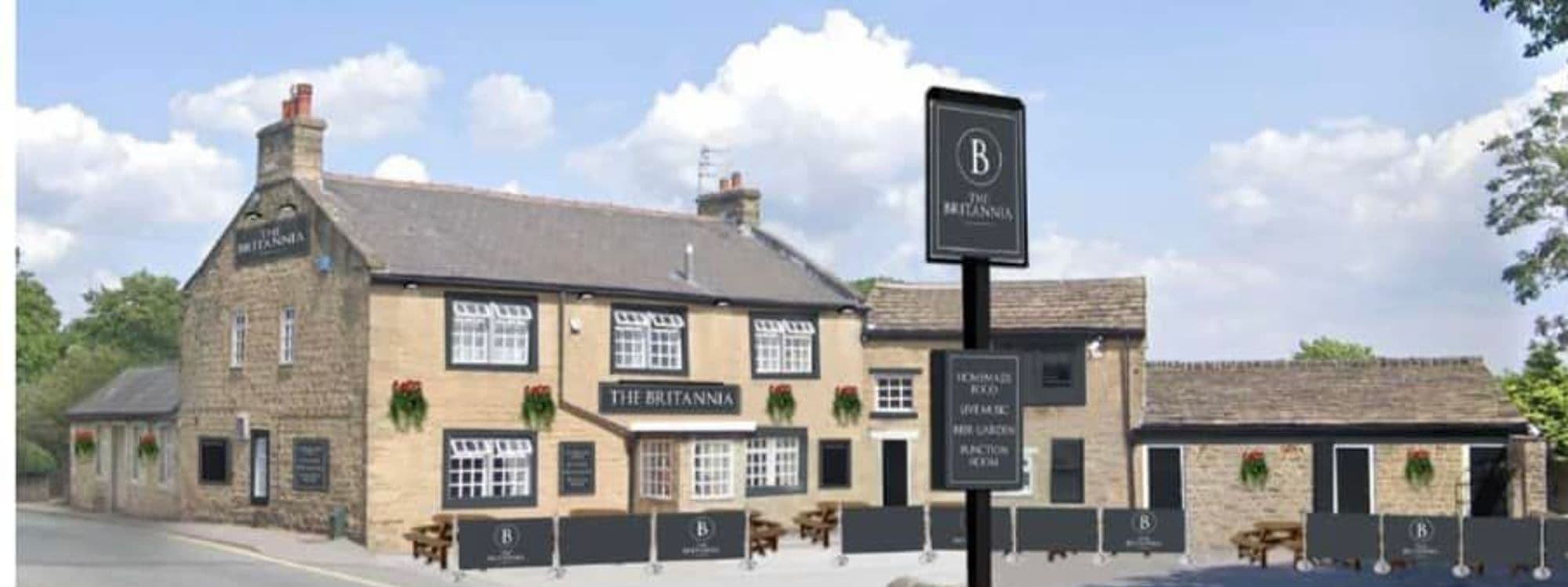 Exterior view of The Britannia, a traditional British pub with stone walls, black window frames, and hanging flower baskets under a clear blue sky. A prominent signboard features the pub's name in front of the building.
