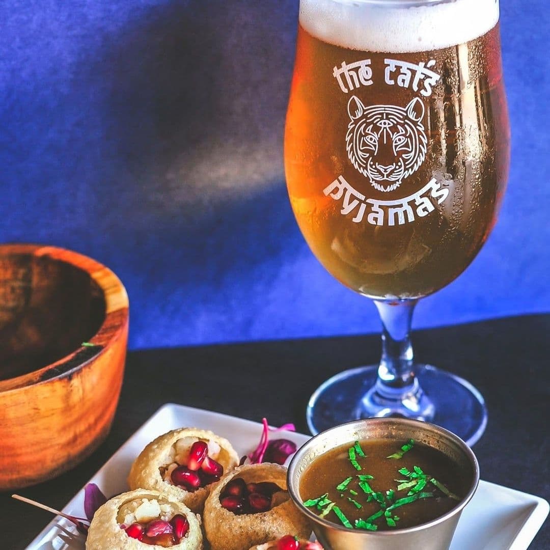 A refreshing beer in a branded glass with 'The Cat's Pyjamas' logo, accompanied by a plate of appetizing Indian snacks, set against a blue backdrop at The Cat’s Pyjamas – Farsley.