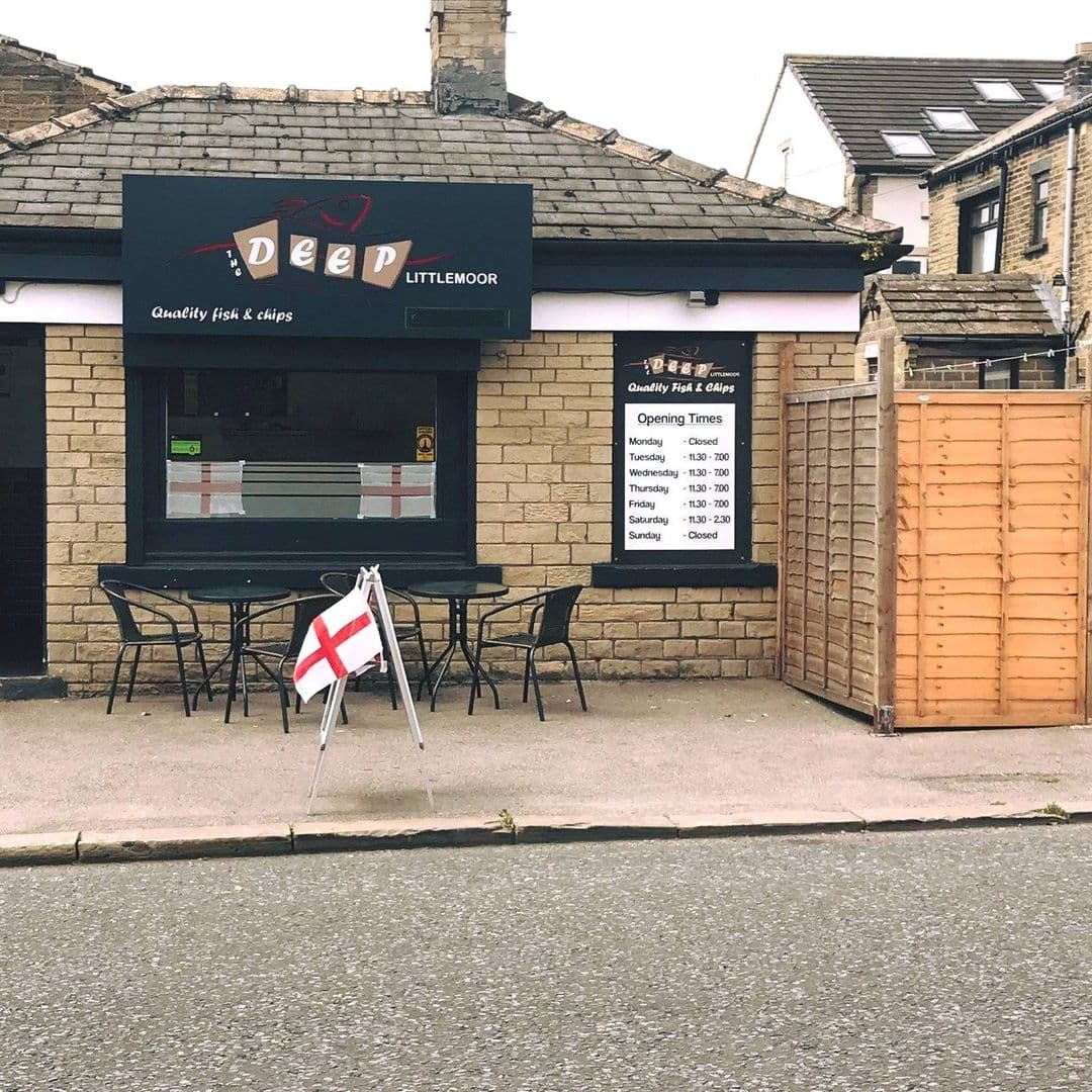 Exterior view of The Deep – Pudsey, a fish and chips shop with a black signboard, outdoor seating, and an English flag. Opening hours are displayed on the window.