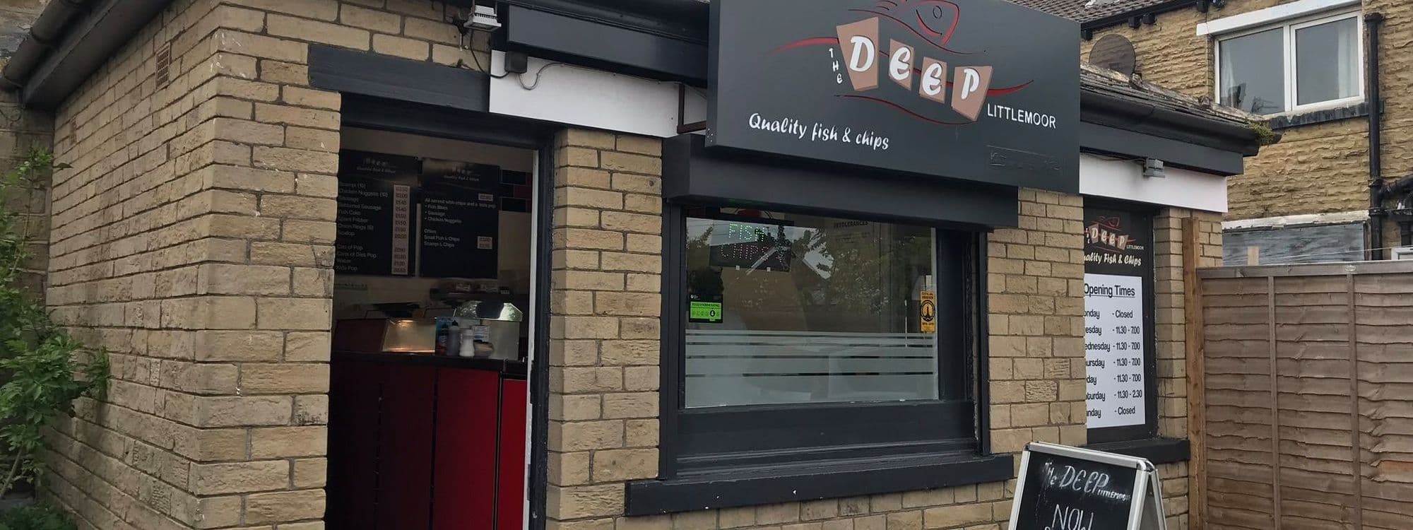 Exterior view of The Deep – Pudsey, featuring a quaint fish and chips shop with a black storefront sign, stone walls, and a visible menu in the window.