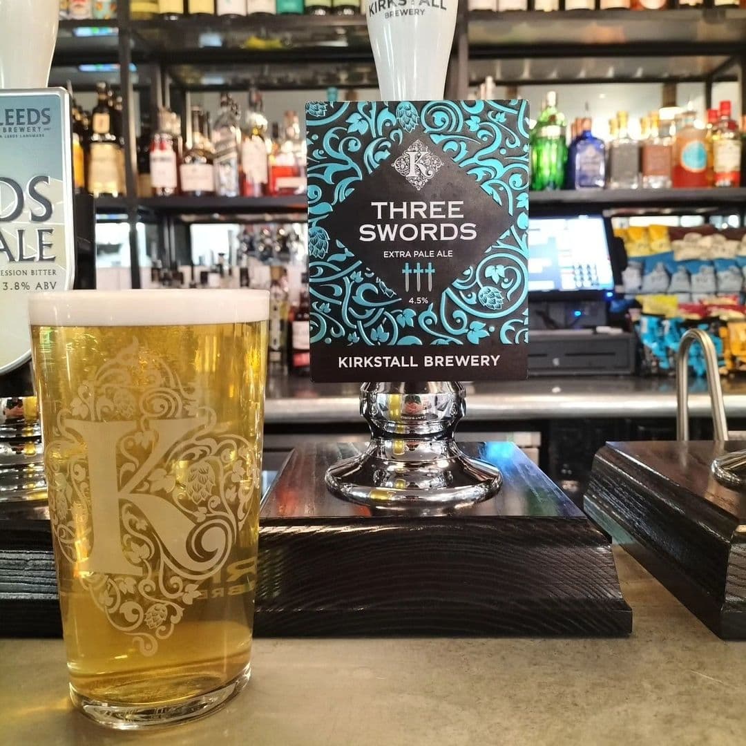 A pint of beer on a bar counter with a decorative glass and a "Three Swords Extra Pale Ale" tap from Kirkstall Brewery in focus, at The Tetley.