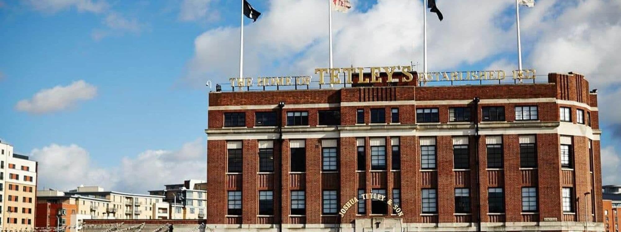 Exterior view of The Tetley, a red brick building with large windows and gold lettering on the facade under a blue sky with scattered clouds.