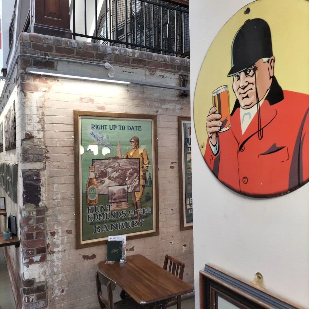 Interior of The Tetley showing vintage advertisements on brick walls, a wooden table with chairs, and a round sign featuring a cartoon man holding a beer.