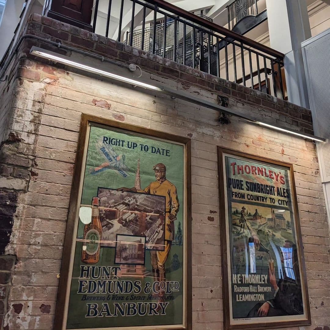 Interior view at The Tetley featuring vintage brewery advertisements on a brick wall, with a metal staircase and balcony in the background.