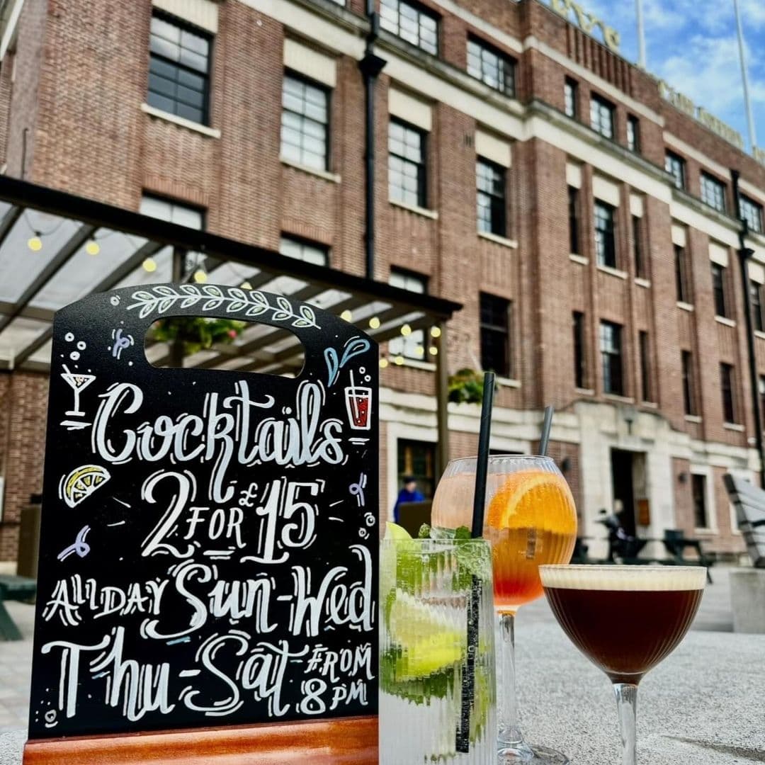 Cocktail promotion sign with "2 for £15" offer in front of The Tetley, with colorful cocktails on a table and the brick facade of the building in the background.