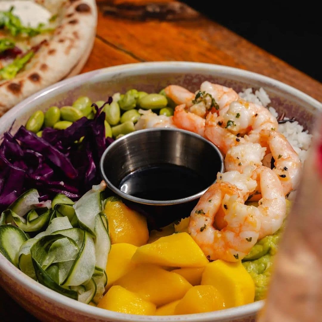 Colorful bowl of food at Treehouse Bar and Kitchen featuring shrimp, mango, edamame, red cabbage, cucumber, and a side of flatbread, with a soy sauce dip in the center.