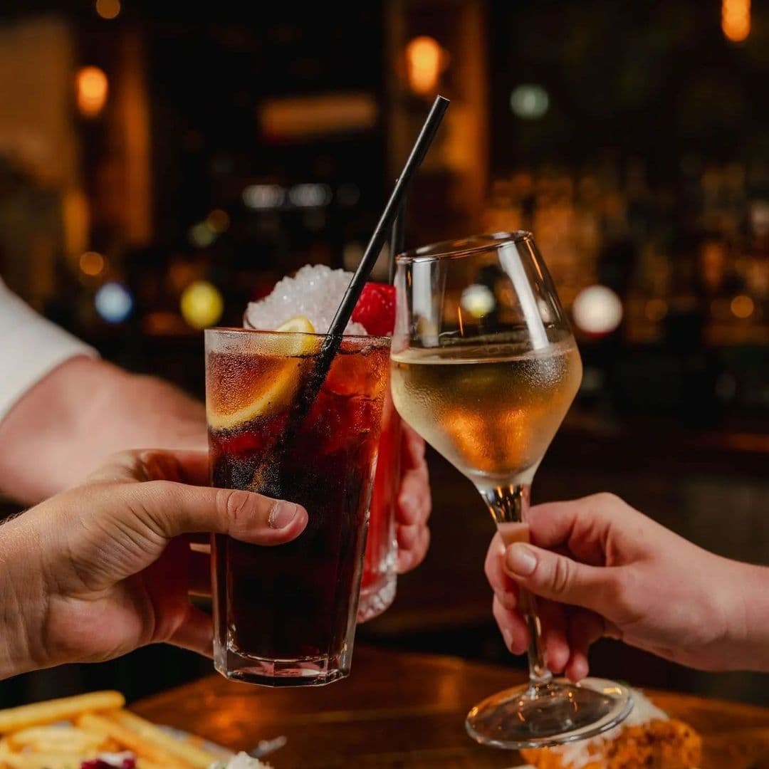 Two people toasting with drinks at Treehouse Bar and Kitchen, one holding a cocktail with a straw and lemon slice, and the other holding a stemmed glass of white wine.