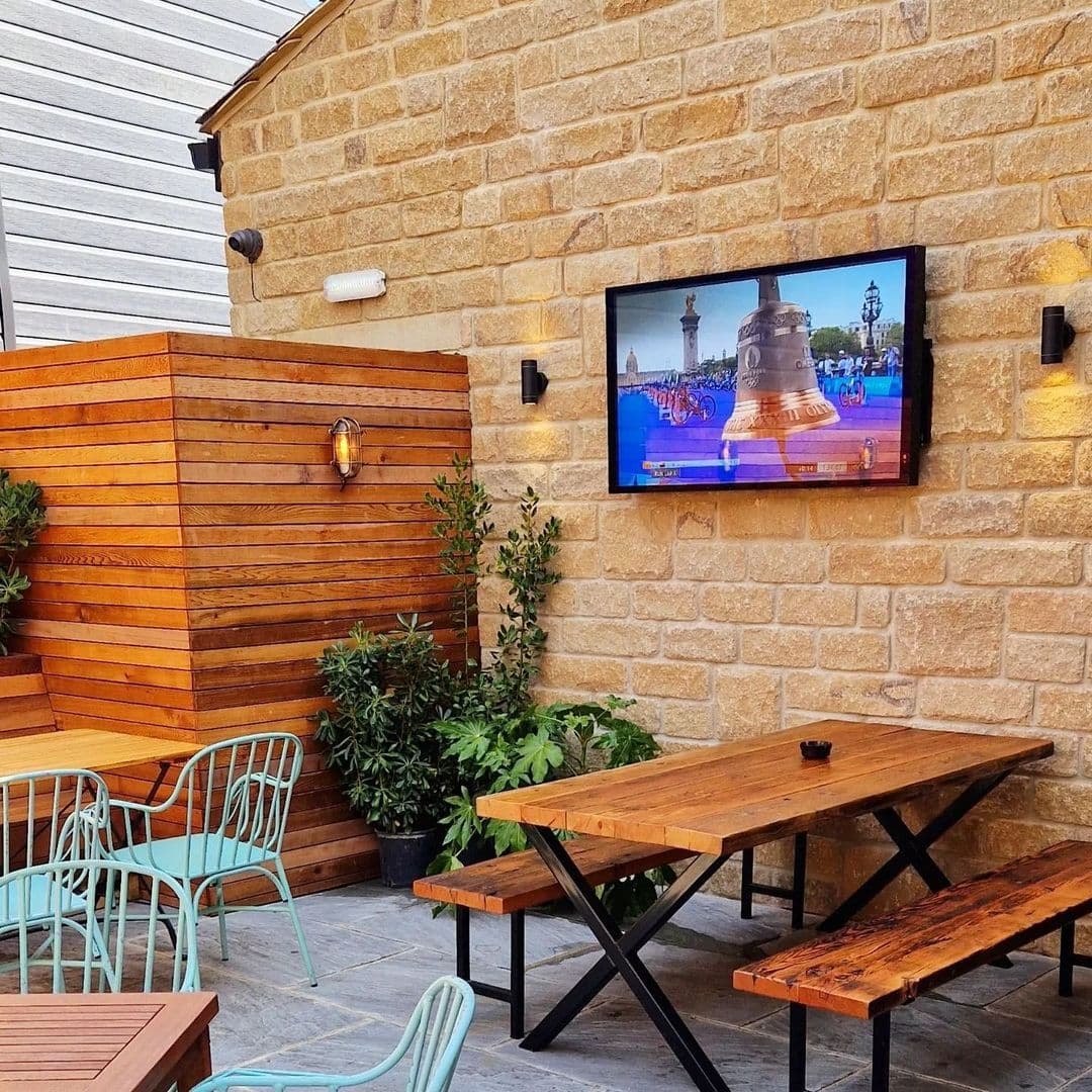 Outdoor seating area at Treehouse Bar and Kitchen with wooden tables and benches, turquoise metal chairs, and a television screen mounted on a stone wall.