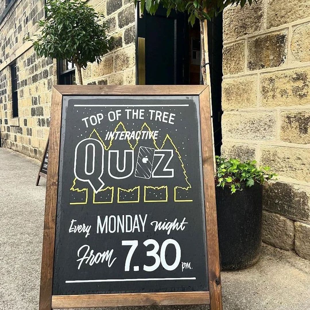 A chalkboard sign outside Treehouse Bar and Kitchen advertising a 'Top of the Tree Interactive Quiz' event every Monday night from 7:30 pm, with a backdrop of a stone wall and a potted plant.