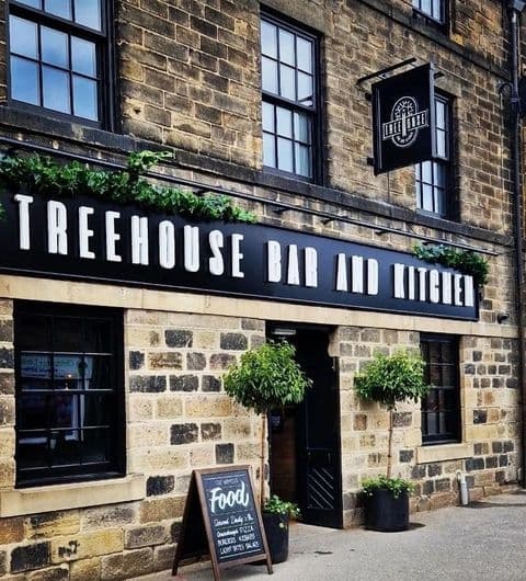 Exterior view of Treehouse Bar and Kitchen with a black awning and signage, stone facade, green plants, and a chalkboard menu.