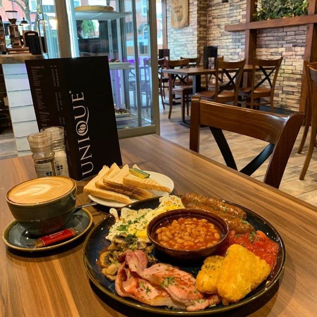 Hearty breakfast plate with eggs, bacon, sausage, beans, and hash browns served at Unique Coffee&Bistro, alongside a cup of cappuccino and toast.