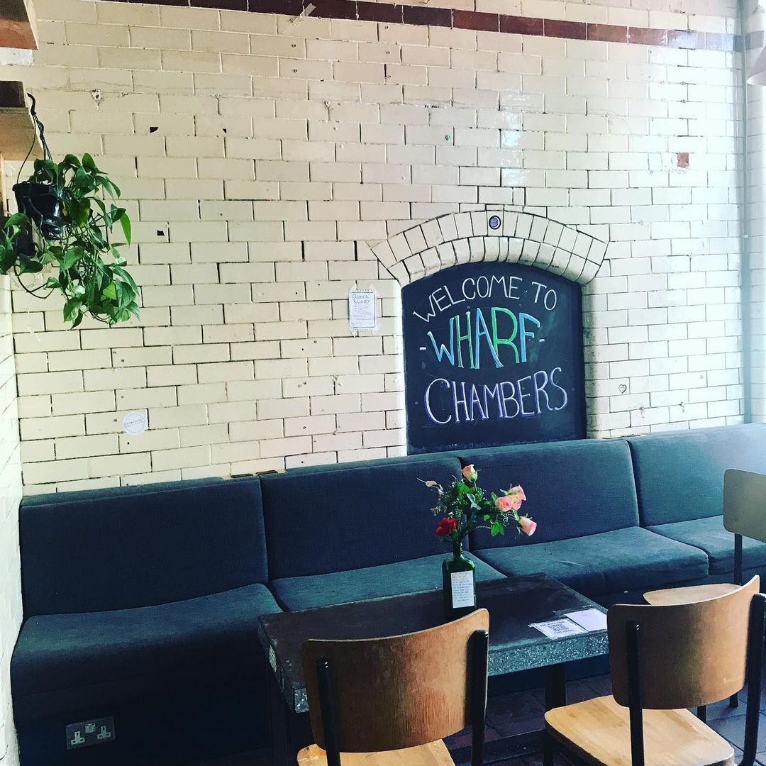 Cozy seating area at Wharf Chambers with a blackboard welcoming visitors, a comfortable dark sofa, wooden chairs, and a brick wall adorned with a hanging plant.