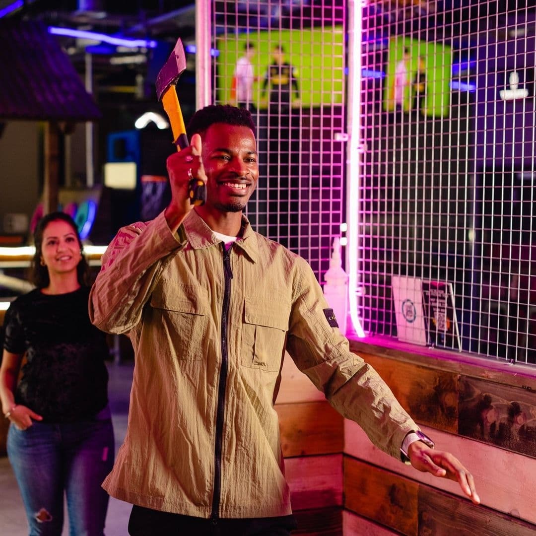 "Smiling man holding an axe at Boom Battle Bar with a woman in the background and colorful neon lights."