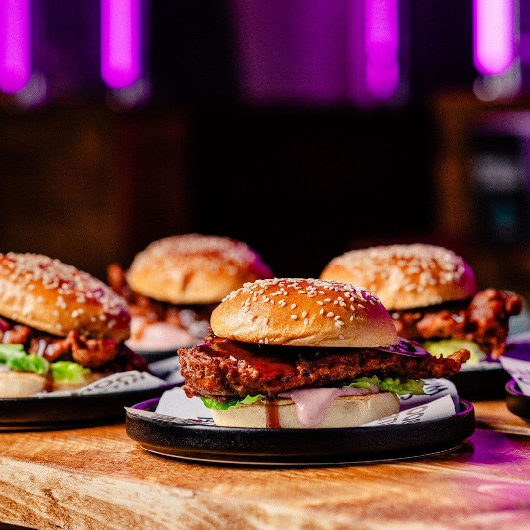Juicy burgers on plates with a vibrant, purple-lit bar atmosphere in the background at Boom Battle Bar.