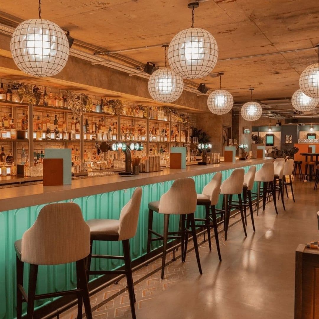 Interior of Electric Shuffle showing a stylish bar with illuminated shelves stocked with various bottles, elegant bar stools in the foreground, and large spherical pendant lights hanging from the ceiling.