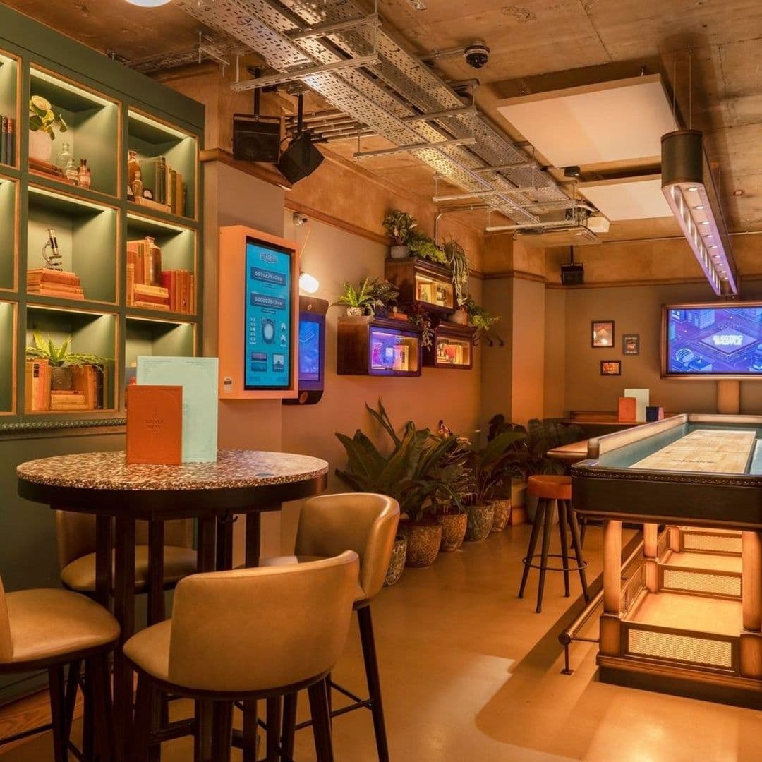 Interior of Electric Shuffle showing a cozy lounge area with bar stools, a shuffleboard table, warm lighting, and greenery.