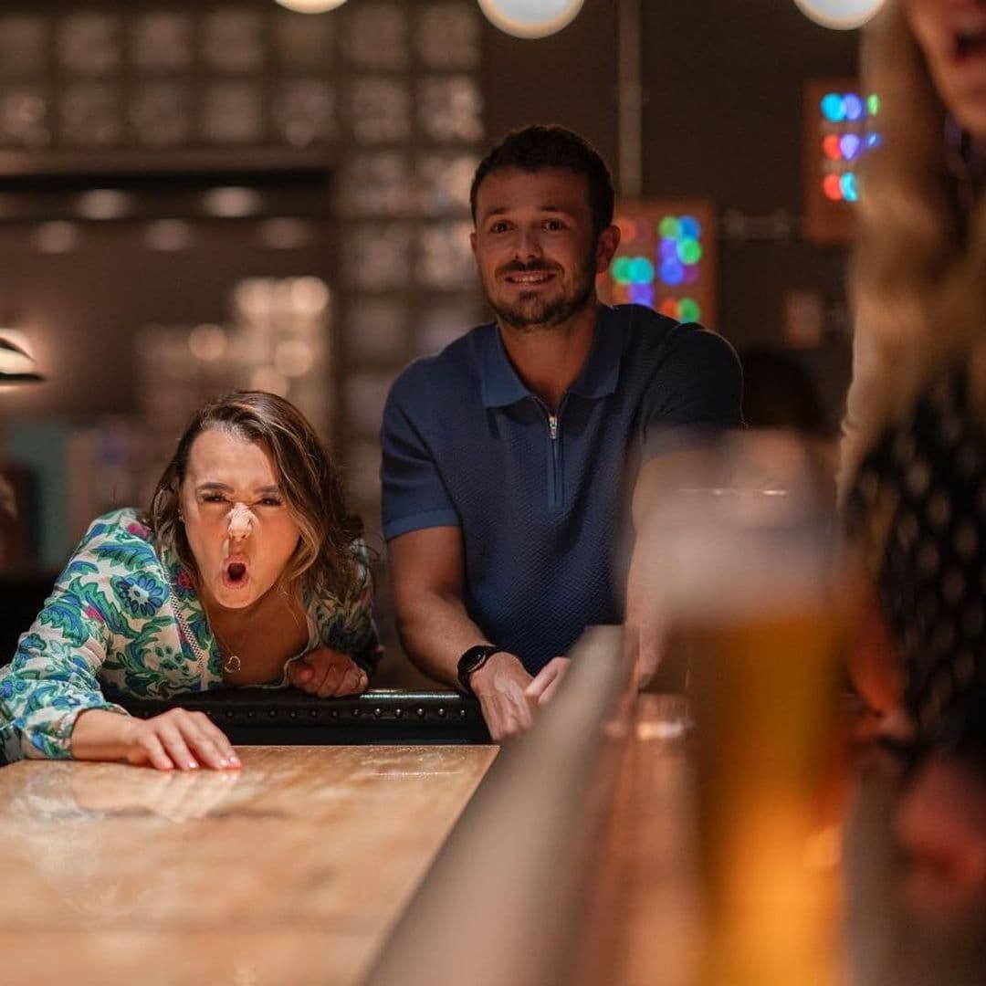 Woman blowing on a shuffleboard puck with excitement at Electric Shuffle while a man watches with a smile, with a pint of beer in the foreground.