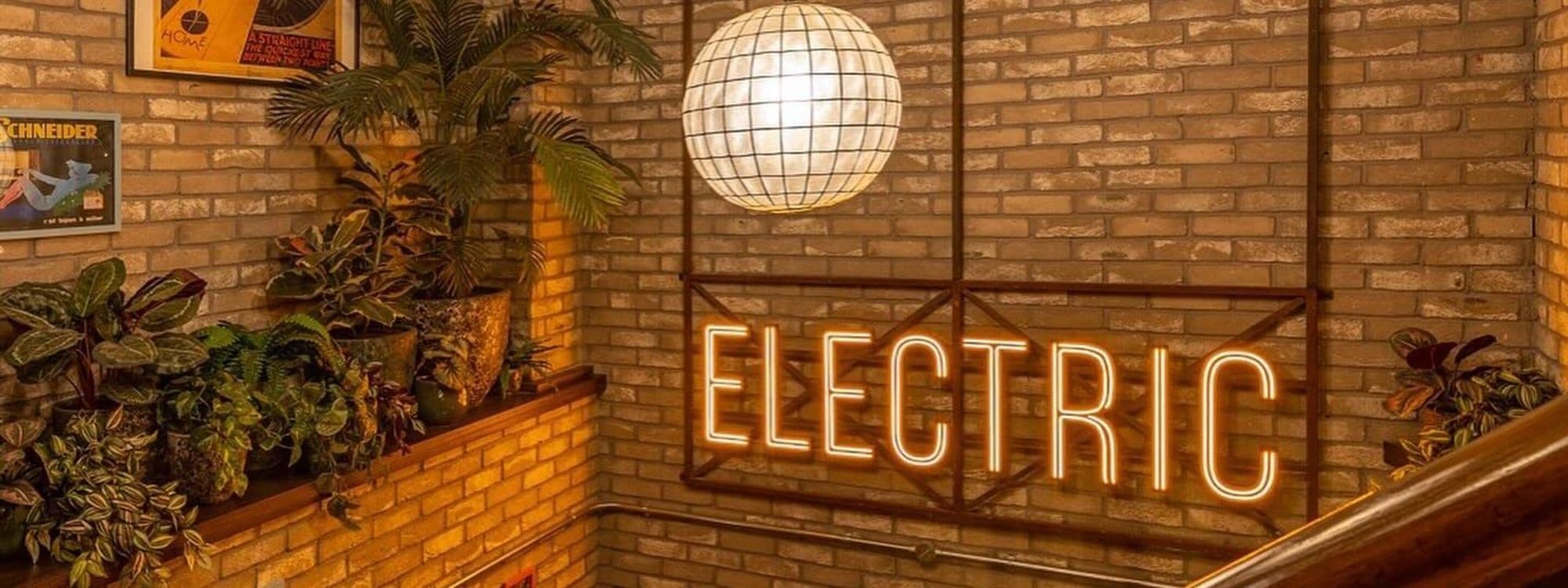 Interior view of Electric Shuffle featuring a neon sign with the word "ELECTRIC" on a brick wall, a large spherical light fixture, and a collection of lush green plants.