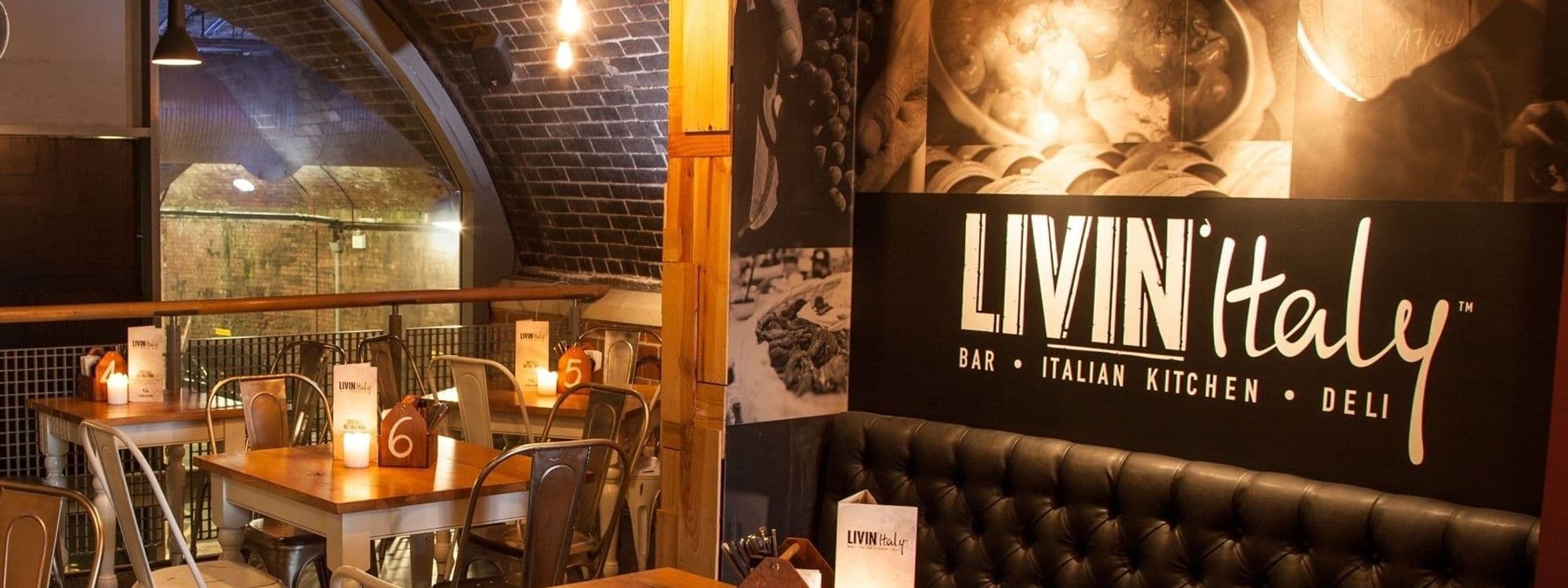 Interior of LIVIN'Italy featuring a cozy bar and dining area with wooden tables, metal chairs, and a large black sofa under warm lighting, with a prominent sign displaying "LIVIN'Italy Bar - Italian Kitchen - Deli".