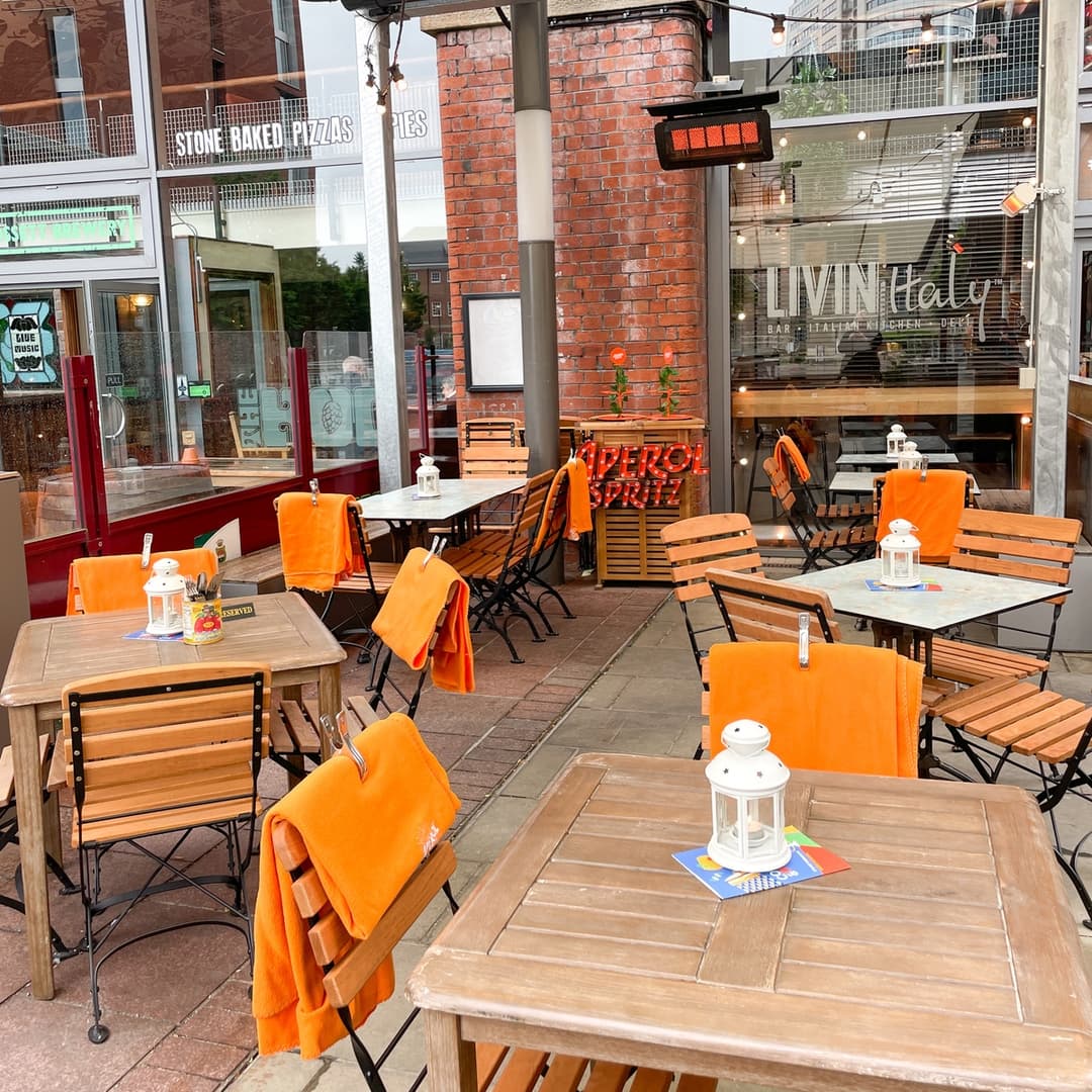 Outdoor seating area at LIVIN'Italy with wooden tables, black chairs, and orange blankets. A lantern and menu on the tables with a view of the restaurant's entrance and signage.