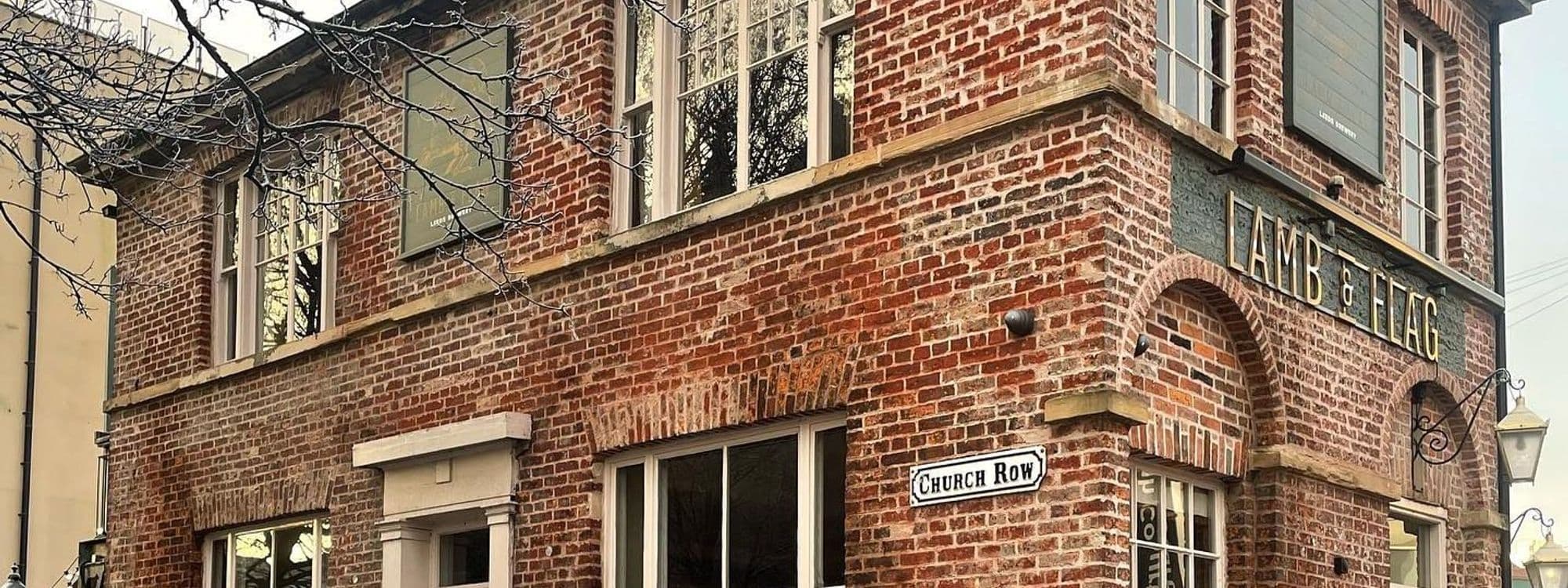 Exterior view of the Lamb and Flag building, a traditional brick structure with white-framed windows and a signboard bearing the name "LAMB & FLAG" above the entrance on Church Row.