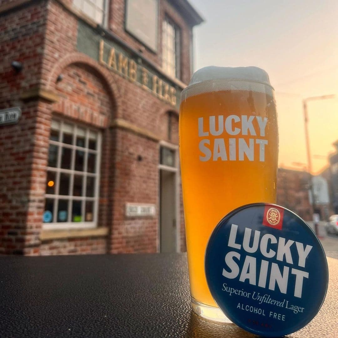 A glass of Lucky Saint unfiltered lager in the foreground with the Lamb and Flag pub sign blurred in the background during sunset.