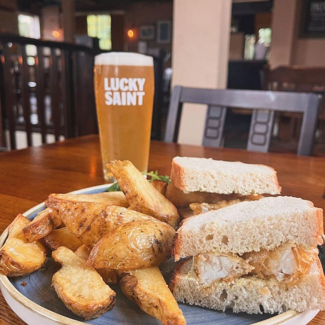A pint of beer with "Lucky Saint" branding next to a plate of fish sandwiches and potato wedges on a table at Lamb And Flag.