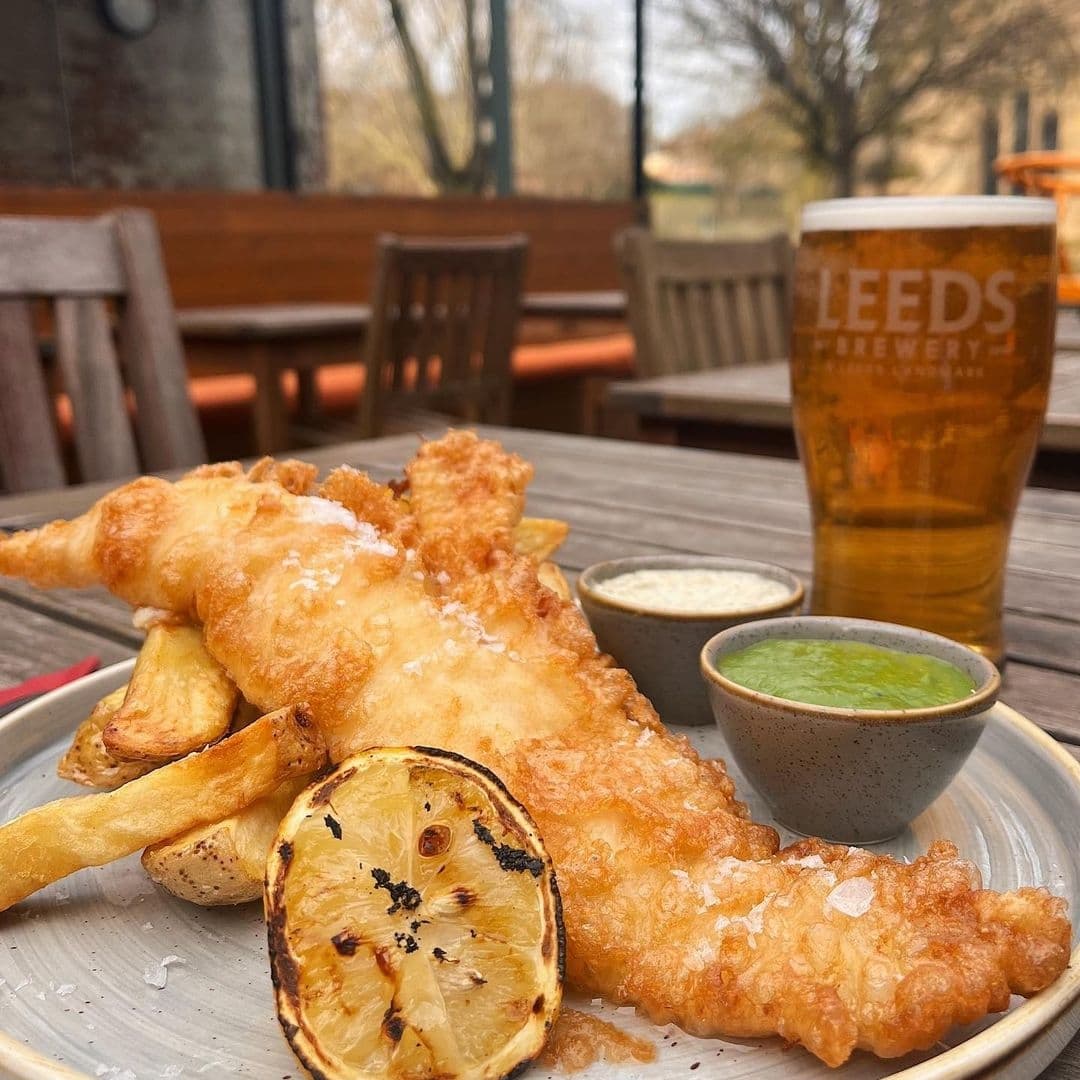 Alt text: "A delicious meal of fish and chips served with a slice of grilled lemon and two dipping sauces, accompanied by a pint of Leeds Brewery beer, at Lamb And Flag."