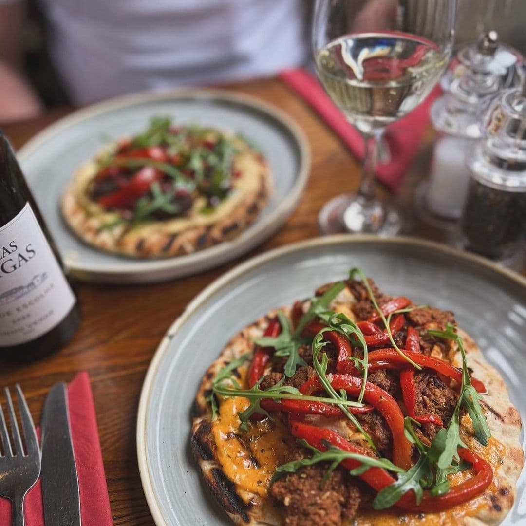Gourmet flatbreads topped with fresh arugula, red peppers, and a drizzle of sauce, accompanied by a glass of white wine and a bottle, at Lamb And Flag.