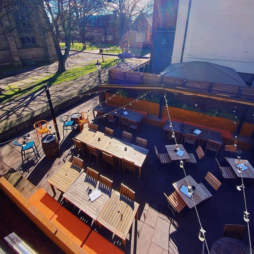 Outdoor seating area at Lamb And Flag with wooden tables and chairs, string lights above, and a view of green trees and a grassy area in the background on a sunny day.