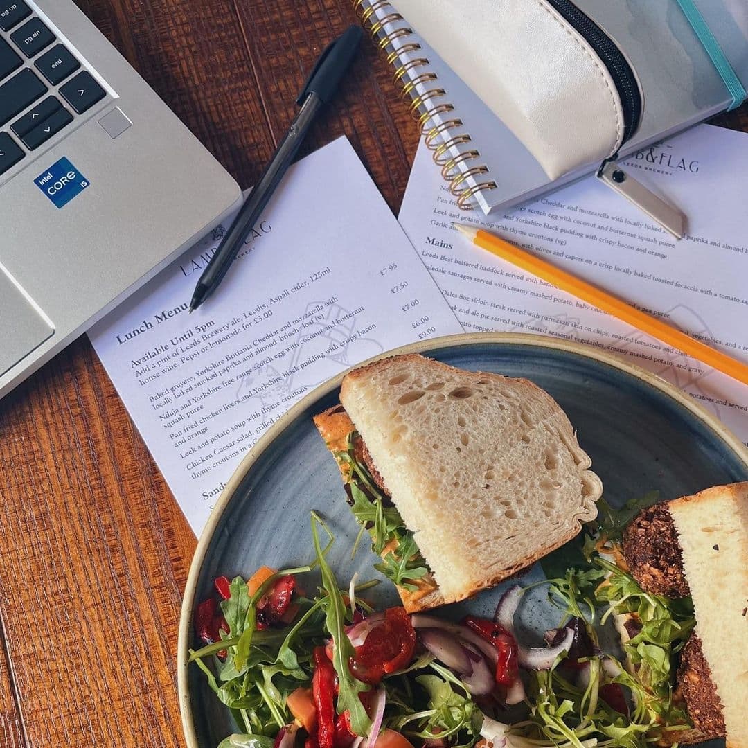 A well-organized work and dining setup at Lamb And Flag featuring a laptop, notepad, pen, and a lunch menu alongside a fresh sandwich with a side salad on a wooden table.