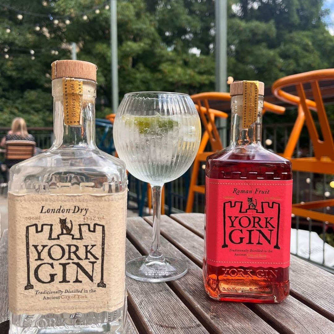 Two bottles of York Gin with distinct labels, one labeled 'London Dry' and the other 'Roman Fruit,' next to a gin tonic glass on a wooden table at Lamb And Flag, with outdoor seating and greenery in the background.