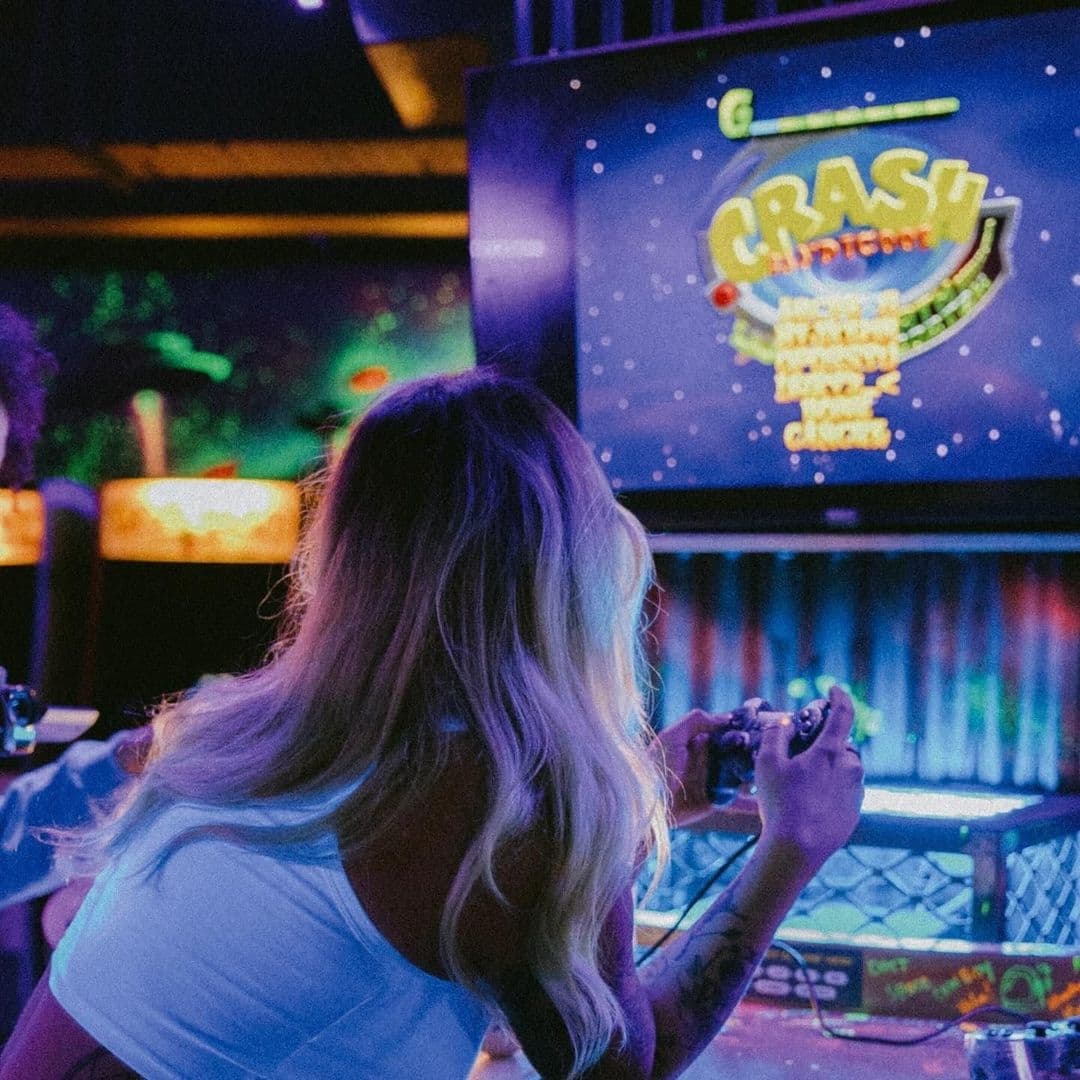 Woman playing a classic video game at NQ64 Arcade Bar, with colorful screen graphics and vibrant atmosphere in the background.