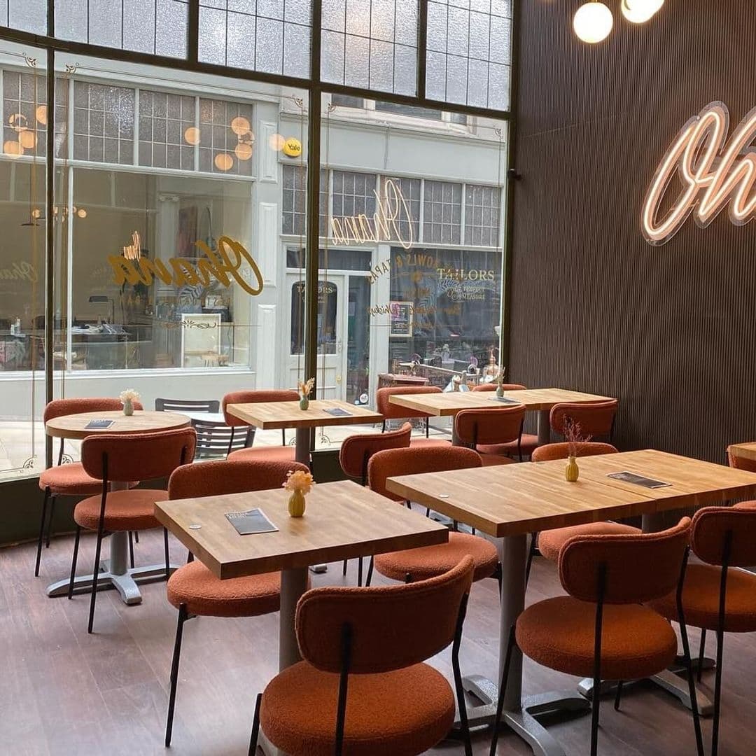 Interior of OHANA cafe with wooden tables, terracotta chairs, and large windows overlooking the street. Decorative lighting and the OHANA logo in neon on the wall add a warm ambiance.