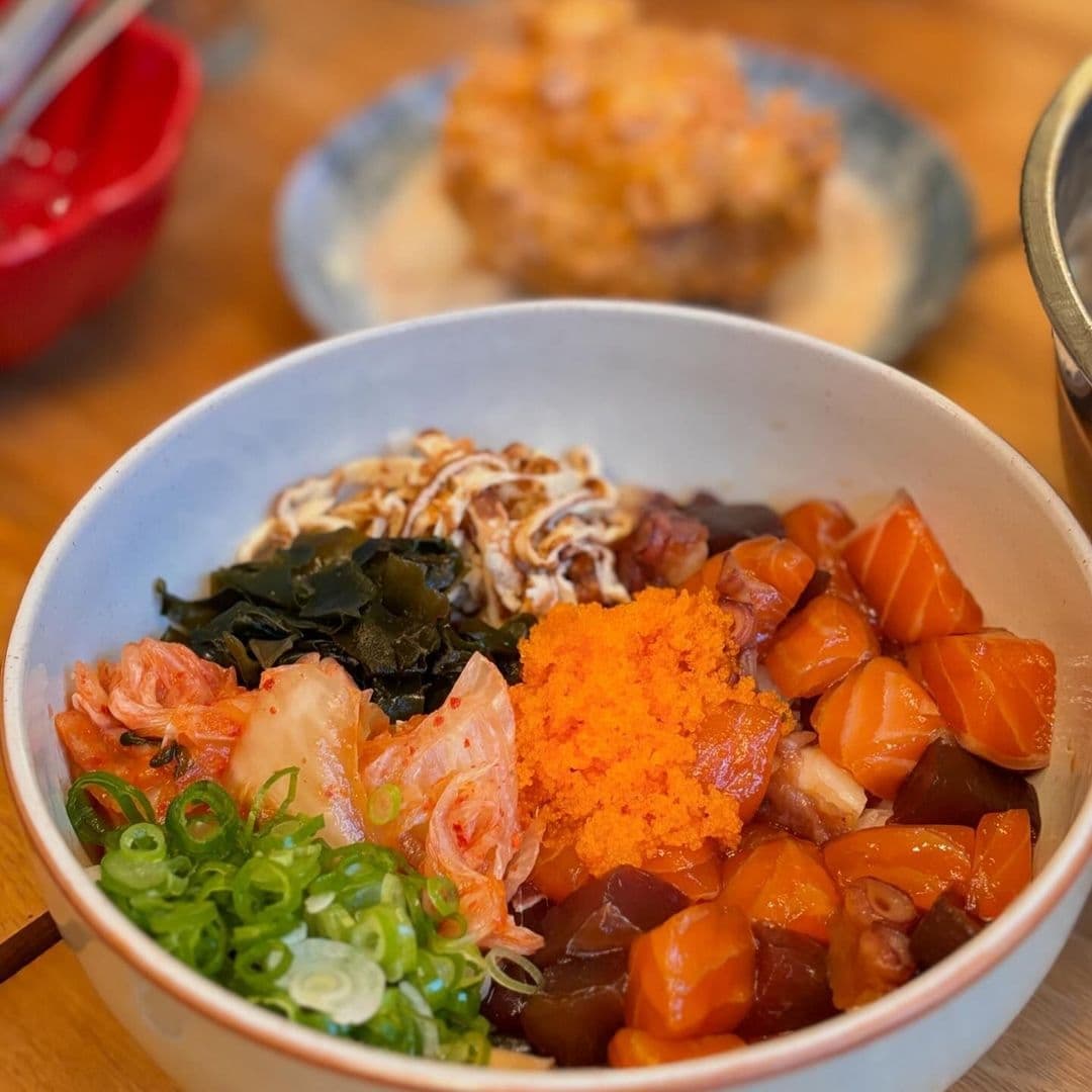 Fresh seafood poke bowl at OHANA featuring diced salmon, tuna, seaweed, green onions, and tobiko, with a side of tempura and miso soup in the background.
