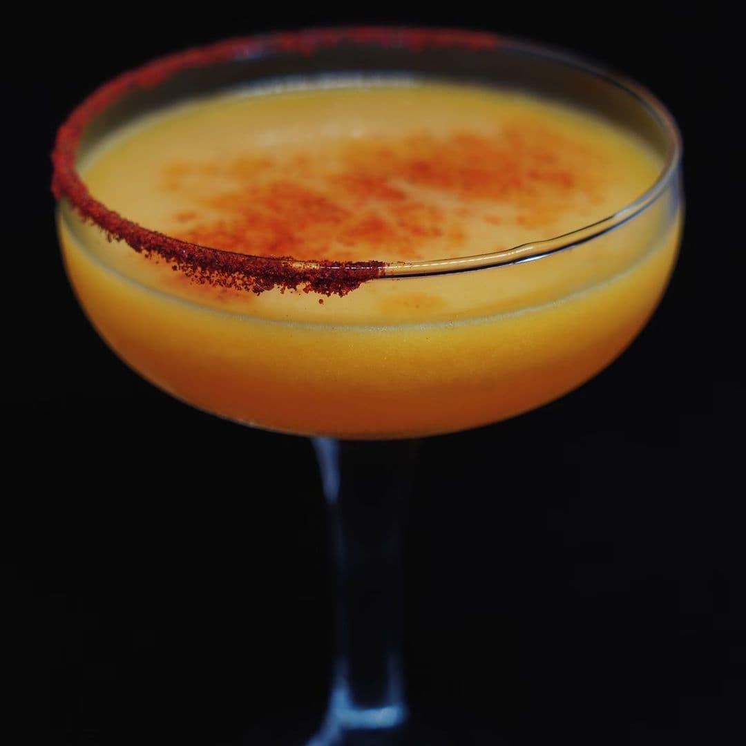 A vibrant orange cocktail with a chili-salted rim, served in a coupe glass against a dark background at OHANA.