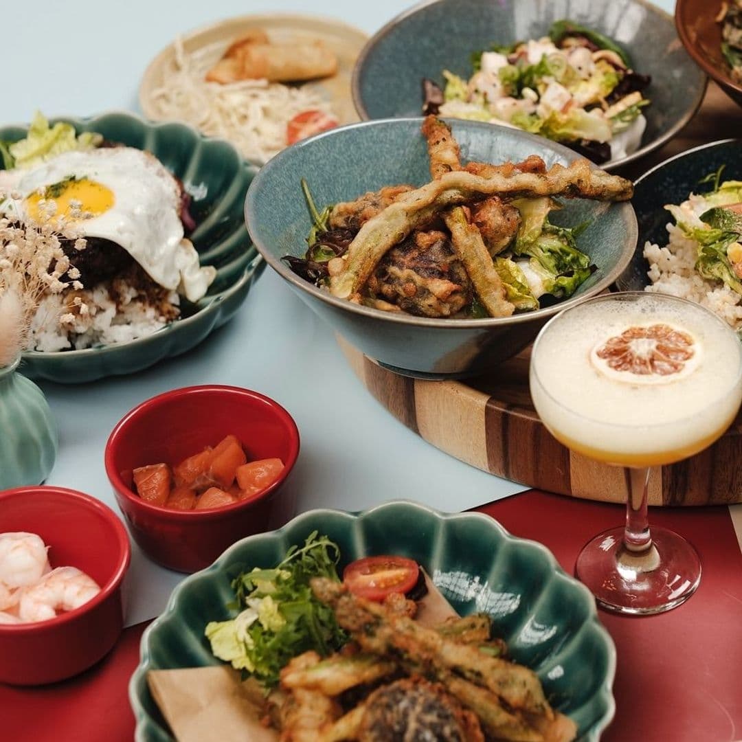 Assorted dishes at OHANA featuring a variety of Asian cuisine, including rice with egg, salad, tempura vegetables, pickled sides, and a frothy cocktail with a citrus garnish.