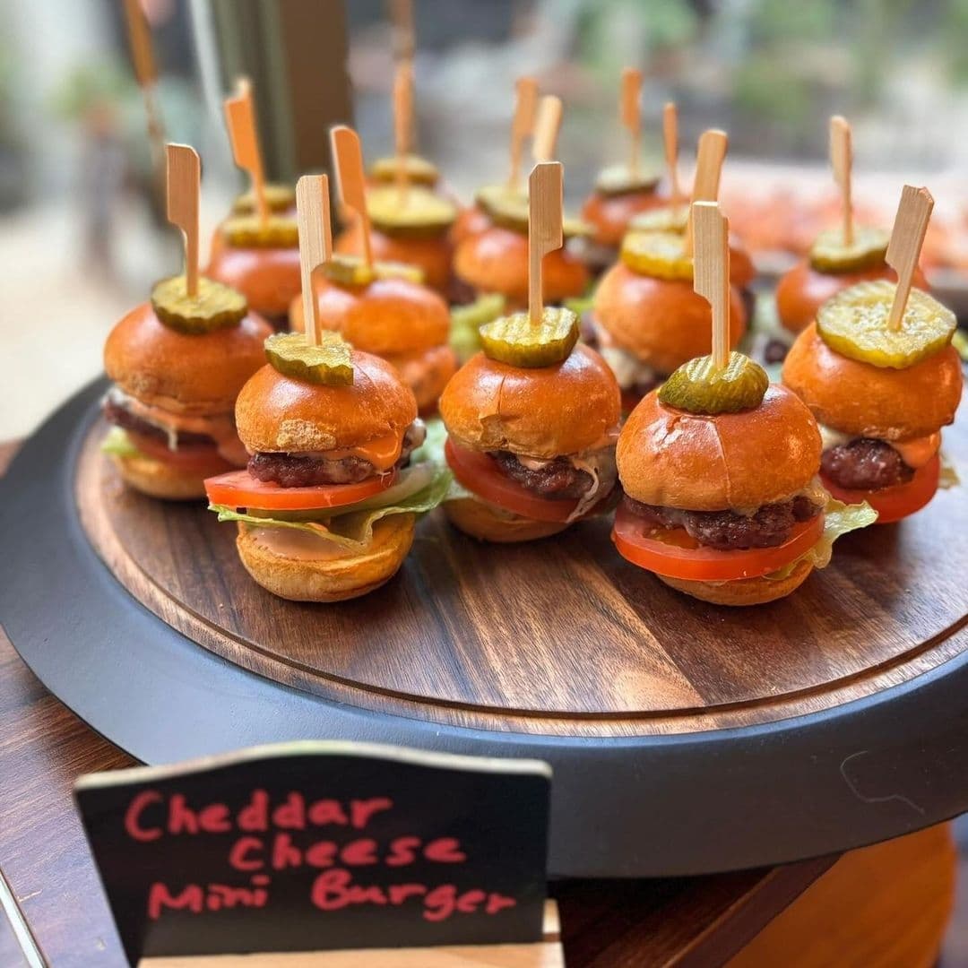 "Appetizing cheddar cheese mini burgers served at OHANA, garnished with pickles on top, presented on a wooden tray."