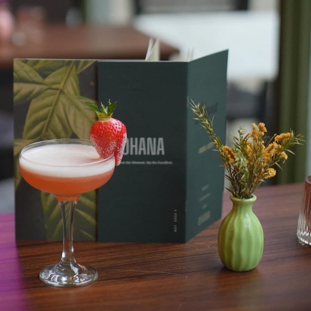 Cocktail with a strawberry garnish in front of an OHANA menu, with a small green vase of yellow flowers on a wooden table.