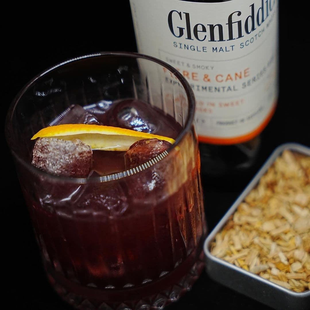 "Close-up of a crafted cocktail with ice and orange peel garnish at OHANA, with a bottle of Glenfiddich single malt scotch and a tin of dried snacks in the background."