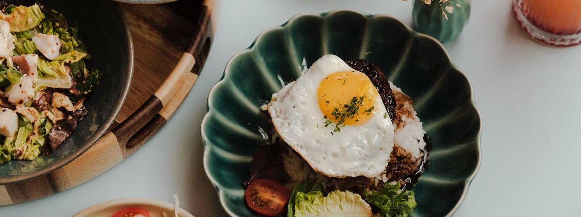 Alt text: "Gourmet dining at OHANA featuring a vibrant dish with a sunny-side-up egg atop seasoned rice, garnished with herbs, alongside a fresh salad and a refreshing beverage in a serene setting."