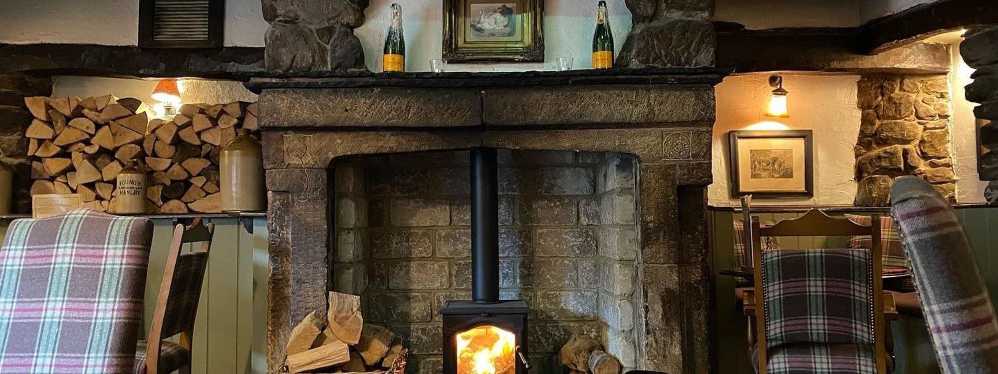 Cozy interior of Scotts Arms with a lit fireplace, stone mantle adorned with firewood, vintage bottles, and framed pictures, flanked by tartan-upholstered chairs.